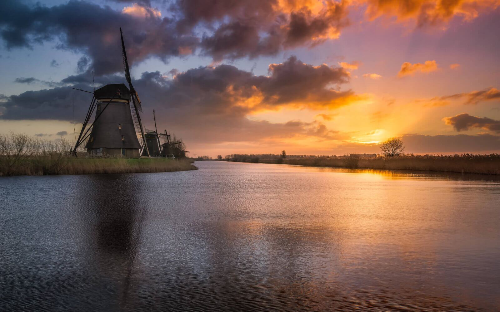 Een kalme, rustige zonsopkomst bij de molens van Kinderdijk. De zon straalt zijn stralen omhoog en kleurt het wolkendek.
